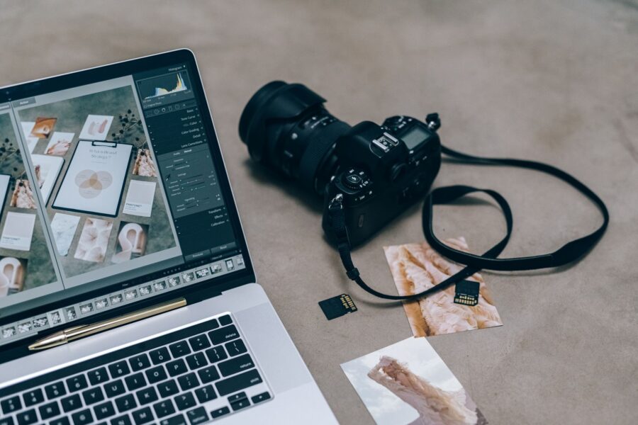 Laptop showing a photo editing program beside a DSLR camera, SD cards, and printed photos on a concrete surface