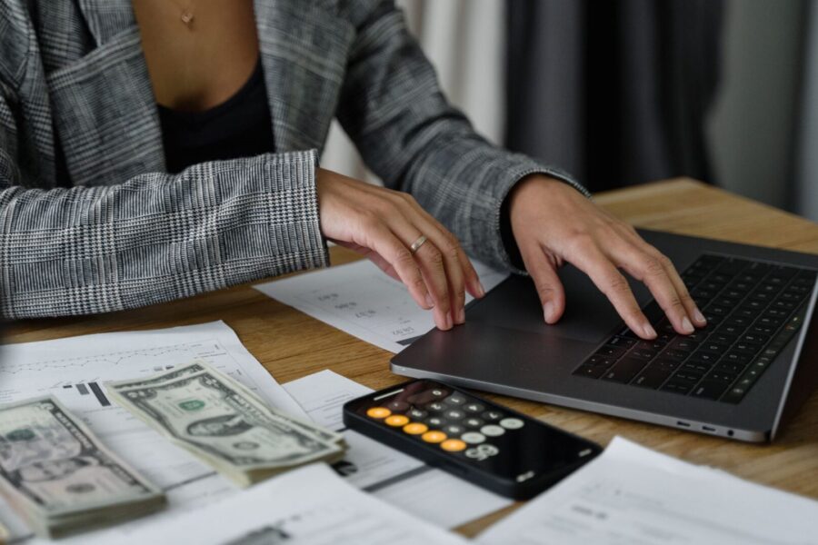 Person using a laptop beside cash, a phone calculator, and financial reports to plan a print-on-demand budget