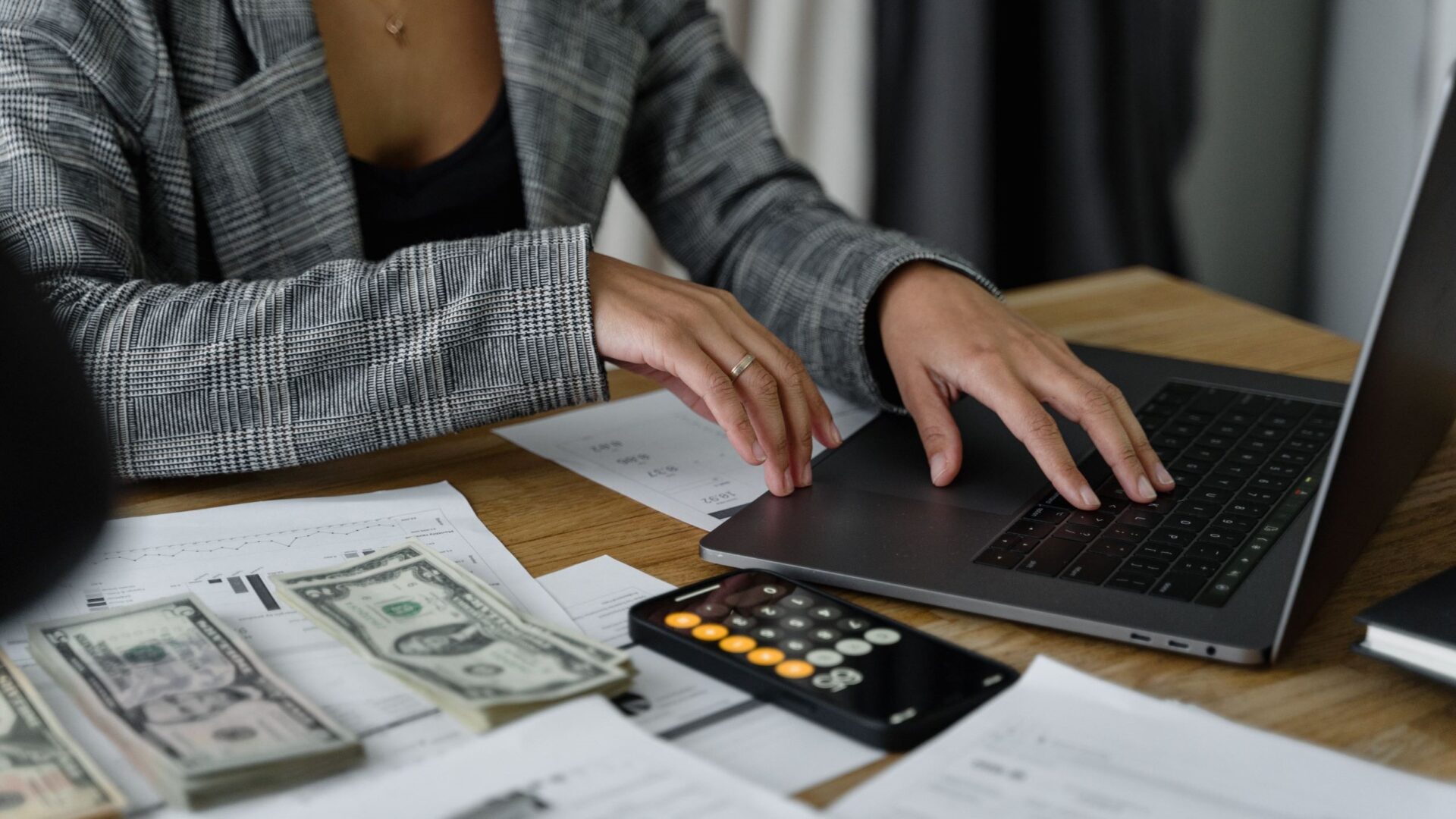 Person using a laptop beside cash, a phone calculator, and financial reports to plan a print-on-demand budget