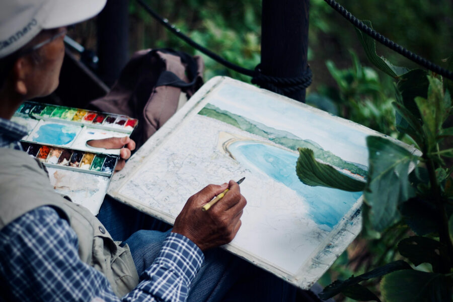 Artist painting a coastal watercolor landscape outdoors, showing the creative process behind the artwork