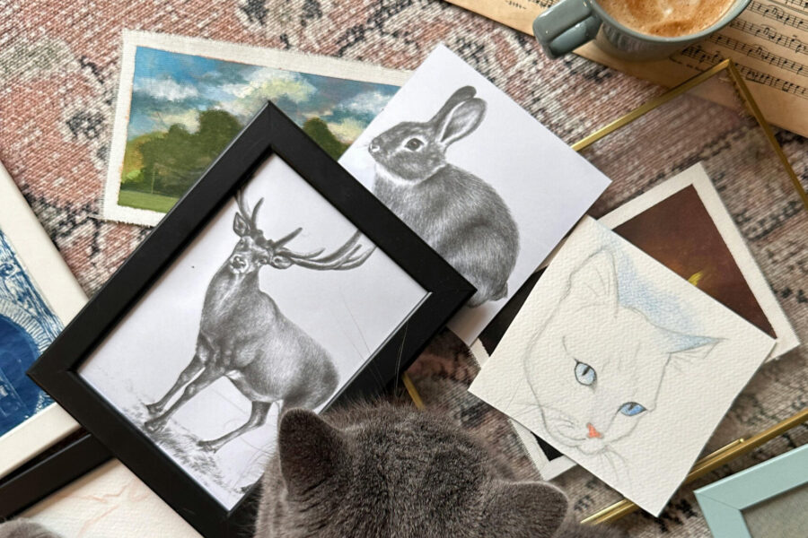 Framed animal artwork and art prints arranged on a table beside a gray cat and coffee cup