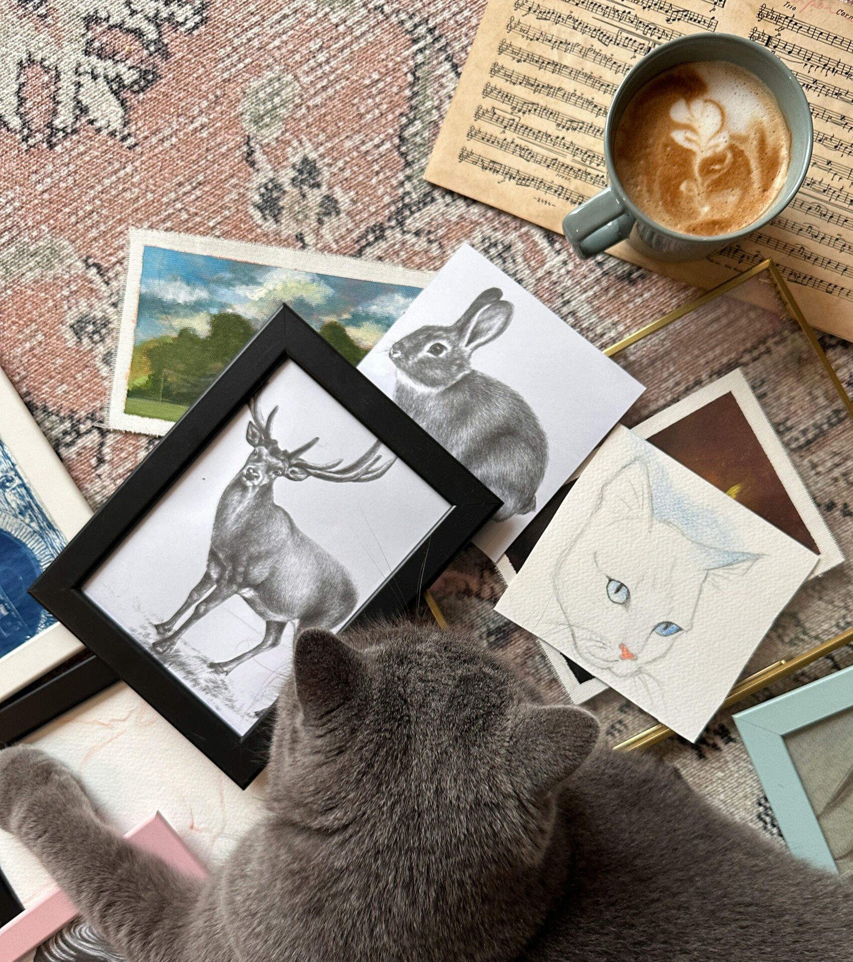 Framed animal artwork and art prints arranged on a table beside a gray cat and coffee cup