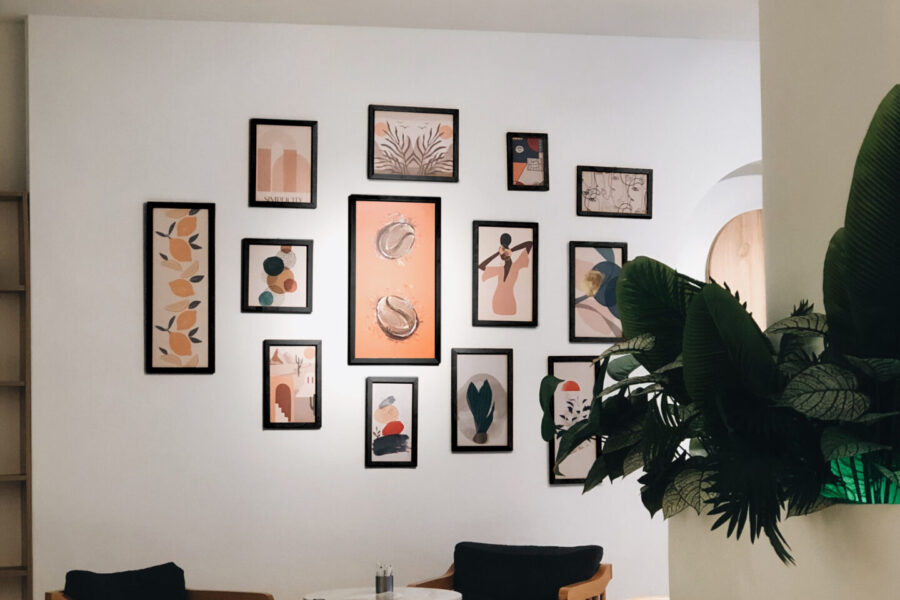 Gallery wall of black-framed limited edition prints in warm terracotta and peach tones above wooden lounge chairs and round white tables
