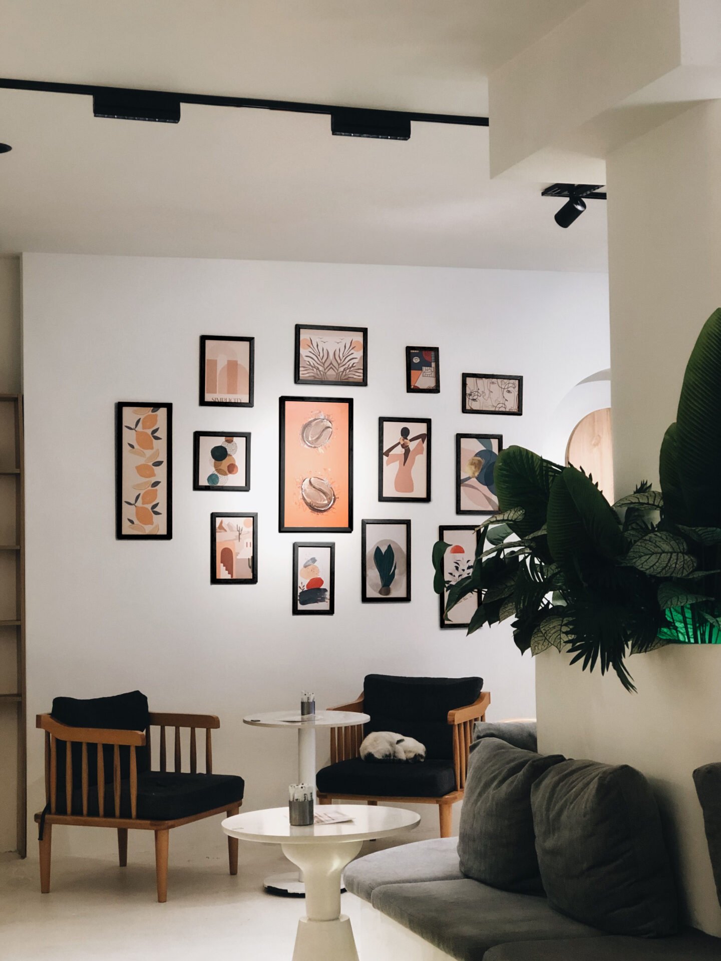 Gallery wall of black-framed limited edition prints in warm terracotta and peach tones above wooden lounge chairs and round white tables