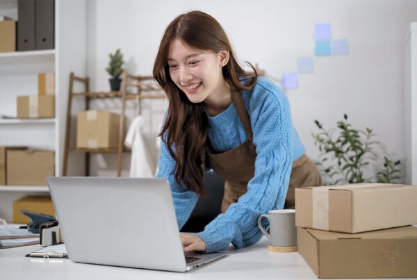 Small business owner using a laptop to manage online store listings with shipping boxes nearby.