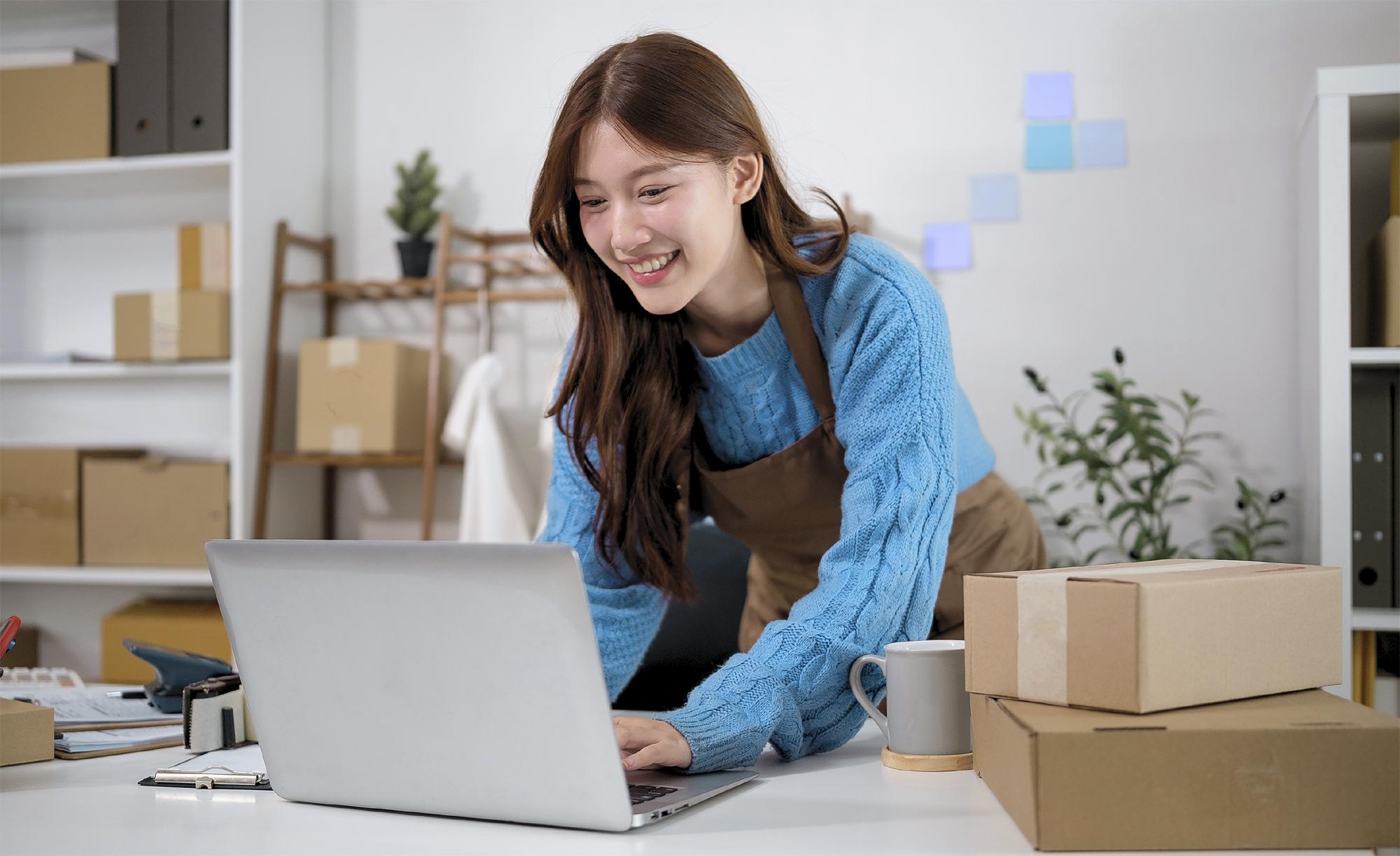 Small business owner using a laptop to manage online store listings with shipping boxes nearby.