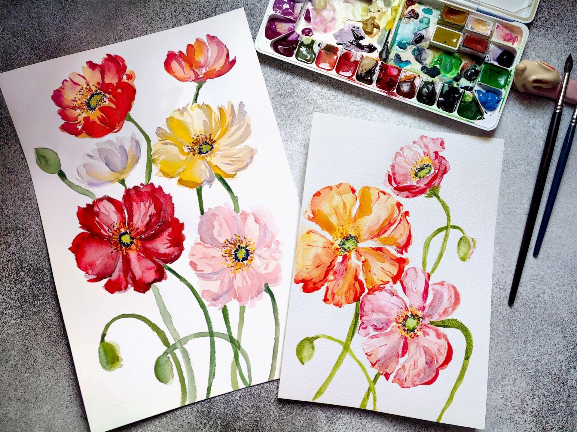 Two textured papers displaying watercolor floral paintings next to art supplies.